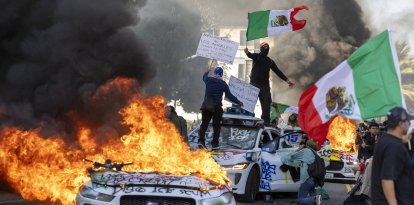 Manifestantes ondean banderas desde lo alto de un coche destrozado, junto a vehículos Waymo en llamas, mientras los manifestantes se enfrentan a las fuerzas del orden en las calles que rodean el edificio federal durante una protesta tras las operaciones federales de inmigración en Los Ángeles, California, el 8 de junio de 2025. Manifestantes incendiaron coches y se enfrentaron a las fuerzas de seguridad en Los Ángeles el 8 de junio, mientras la policía mantenía a los manifestantes alejados de las tropas de la Guardia Nacional que el presidente Donald Trump envió a las calles de la segunda ciudad más grande de EE.UU.(Foto de RINGO CHIU / AFP)
