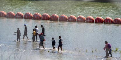 Crisis migratoria: Migrantes caminan junto a una cadena de boyas colocadas en el agua a lo largo de la frontera del Río Grande con México en Eagle Pass, Texas, el 15 de julio de 2023