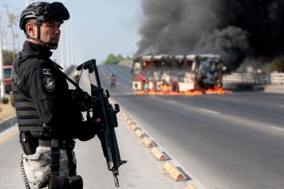 A member of the Prosecutor's Office stands guard near a burning bus at one of the main avenues after it was set on fire by organised crime groups in response to an operation in Jalisco to arrest a high-priority security target in Zapopan, state of Jalisco, Mexico, on February 22, 2026. Armed civilians blocked several roads in the state of Jalisco, in western Mexico, following an operation by federal forces in the town of Tapalpa, local authorities reported. Jalisco, which will host four matches of the upcoming 2026 World Cup, is home to the powerful Jalisco New Generation Cartel (CJNG), and has been rocked by several episodes of violence in recent years. (Photo by Ulises Ruiz / AFP)