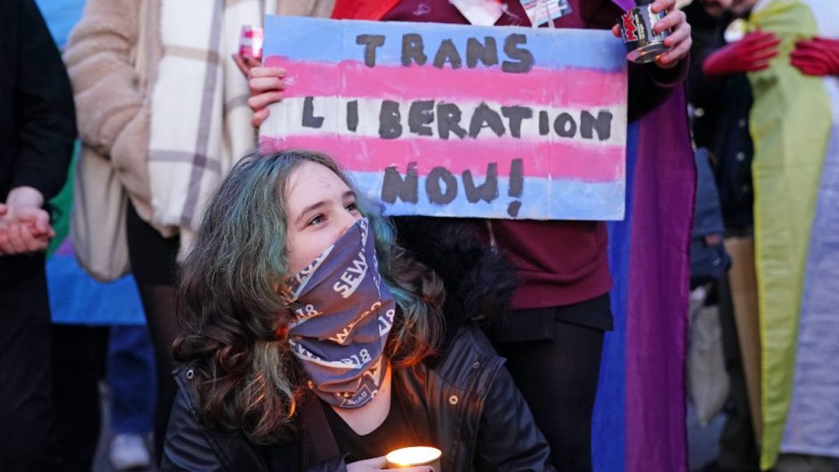 Activista trans con la cara cubierta por una bandana sostiene una vela delante de un cartel que reza 