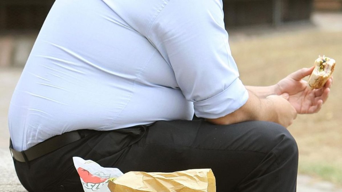 Torzo y piernas de un hombre con sobrepeso mientras come una hamburguesa sentado en un parque.