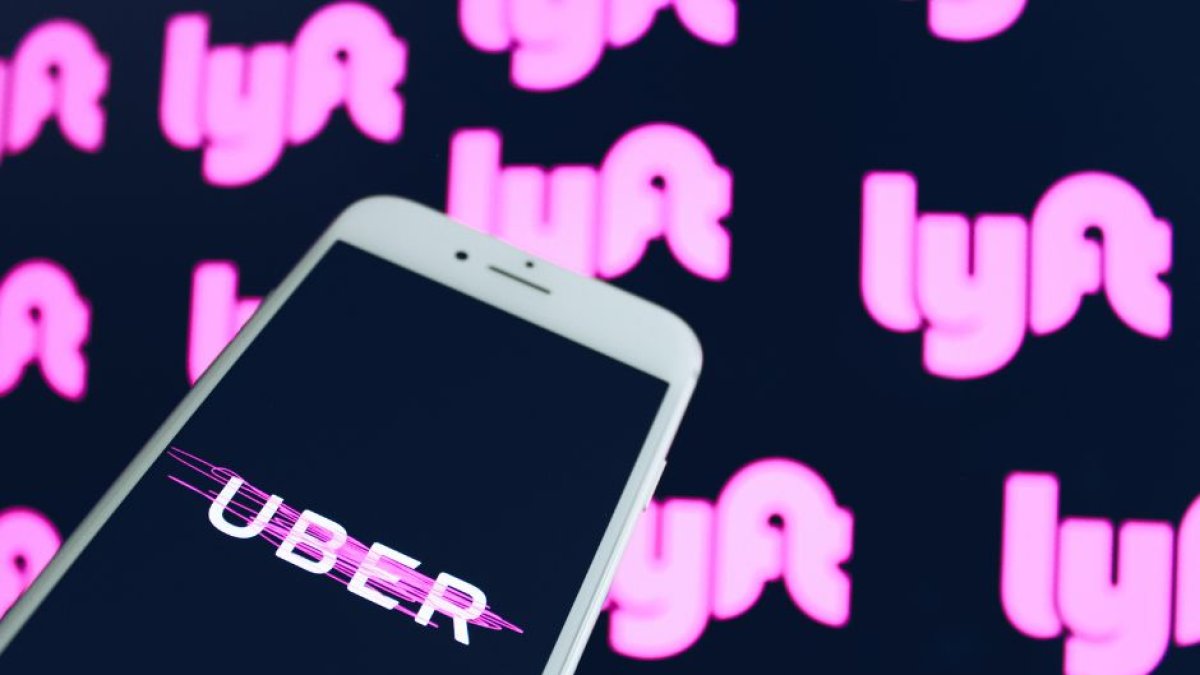 Mano sosteniendo un smartphone con el logo de Uber. Detrás un fondo de pantalla con varios logos de Lyft. Ambas compañías dejarán de operar en Minneapolis a partir del 1 de mayo.