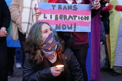 Activista trans con la cara cubierta por una bandana sostiene una vela delante de un cartel que reza 