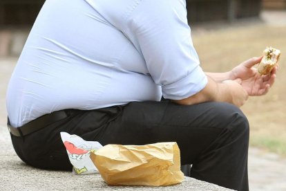 Torzo y piernas de un hombre con sobrepeso mientras come una hamburguesa sentado en un parque.