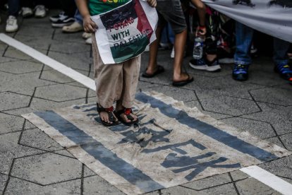 Un niño pisa una bandera con los colores de Israel mientras sostiene un cartel pro Palestina.