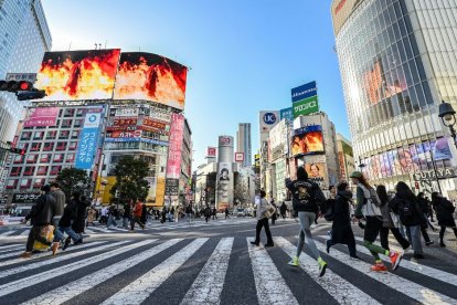 Unos peatones caminan por el cruce de Shibuya mientras un vídeo promocional (arriba) con imágenes de 