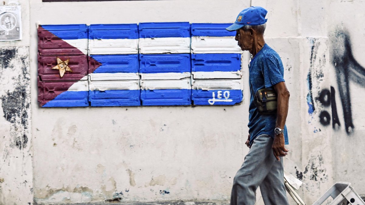 An elderly man walks in front of a mural with the Cuban flag.