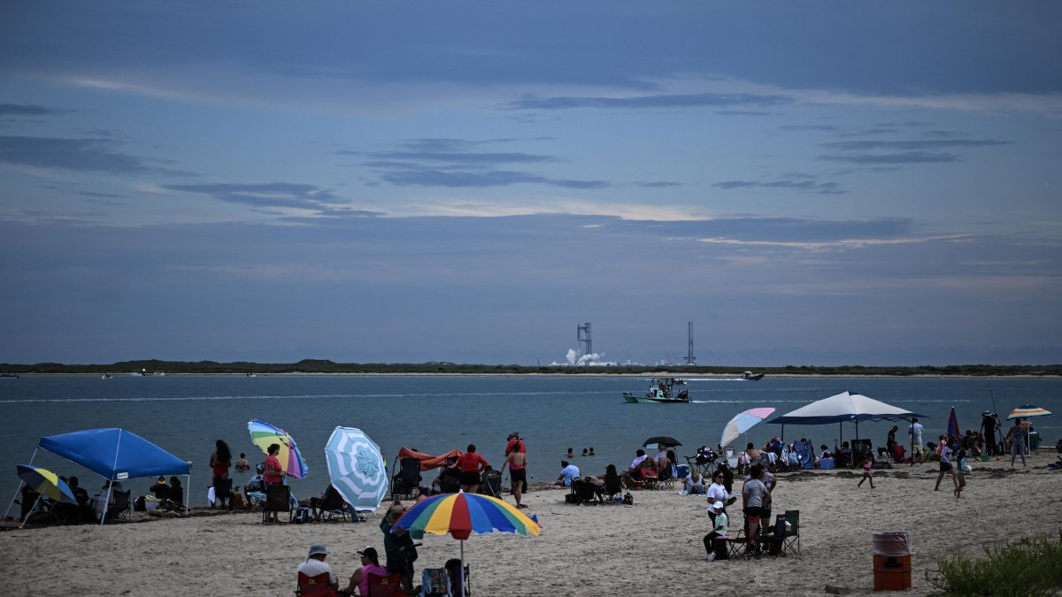 People sit on the beach at South Padre Island, Texas, with SpaceX's Starship spacecraft visible in the background.